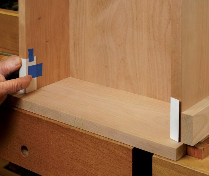 Prep the case. To create the right clearance behind the door (11⁄6464 in.), Gochnour taped cardboard to the front edges of the case. To make the door flush with the hinge side of the case, he clamped on a block there.